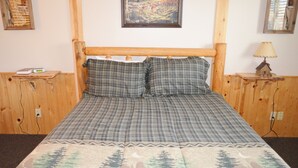 Ein Bett mit karierter Bettdecke und zwei Kissen, ein Holz-Kopfbrett, ein Nachttisch mit Lampe und ein Bild an der Wand.