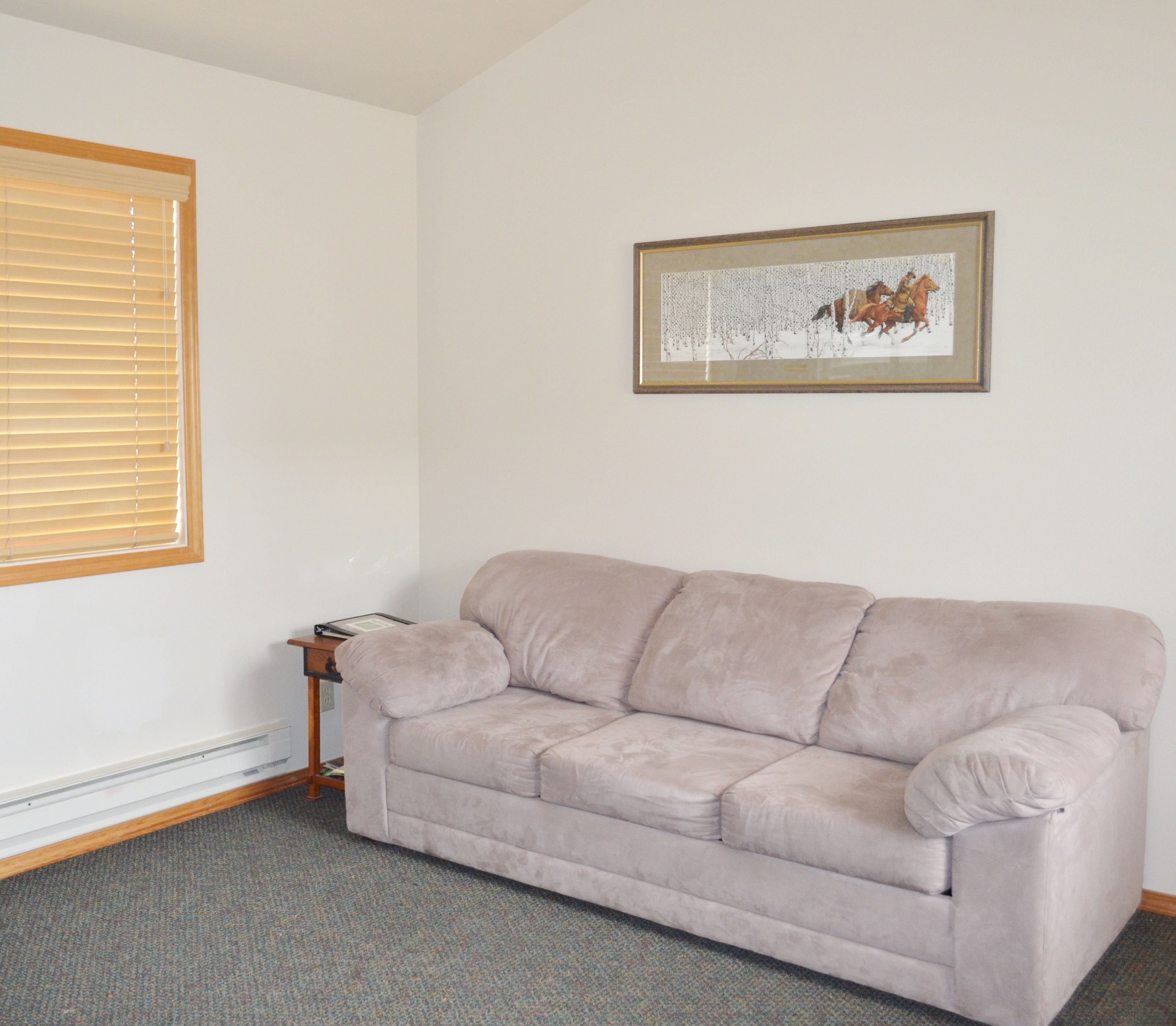 A room with a beige sofa, a framed picture on the wall, a window with blinds, and a small table with a lamp.