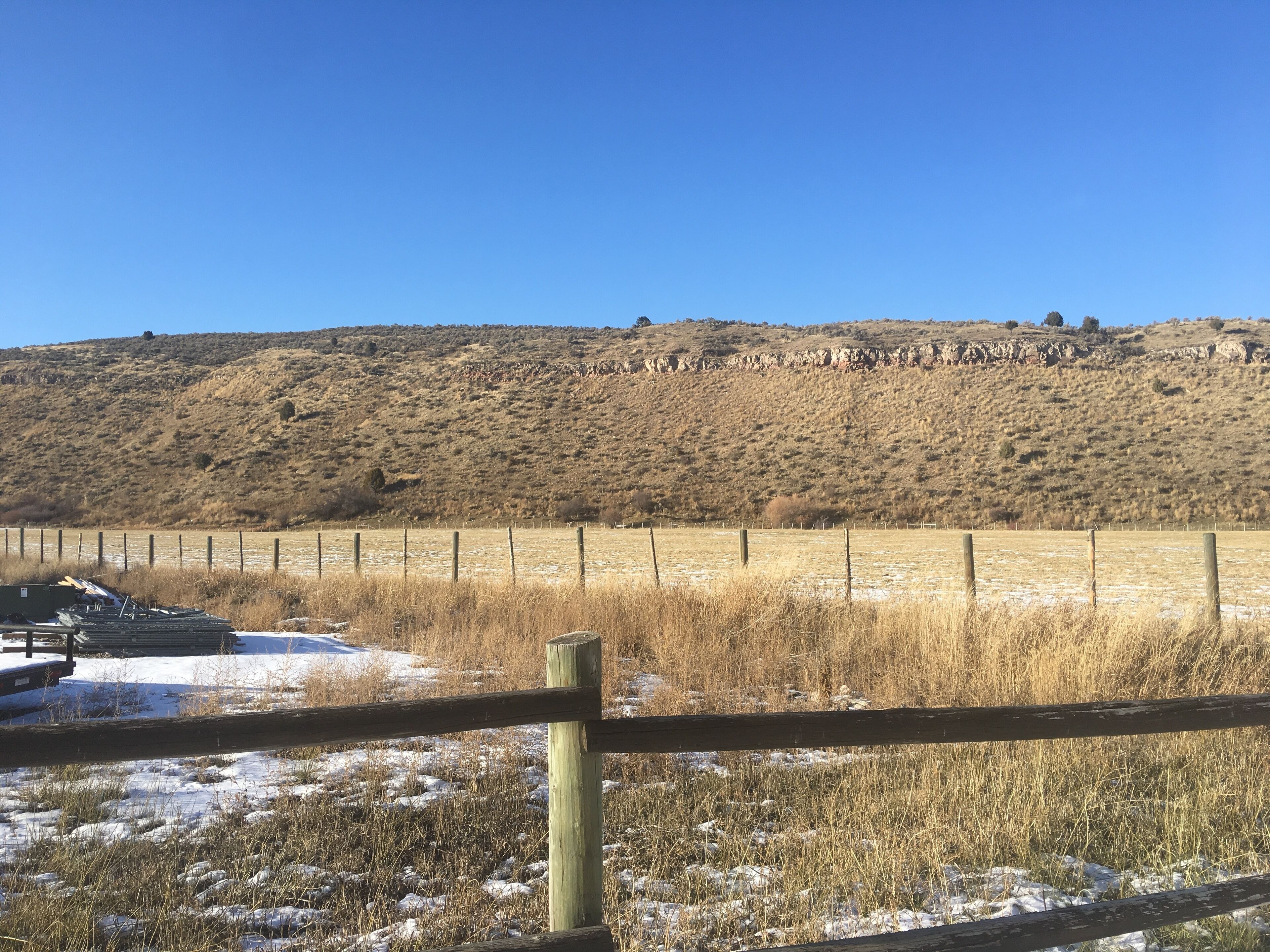 A fenced field with dry grass and a rocky hill in the background.