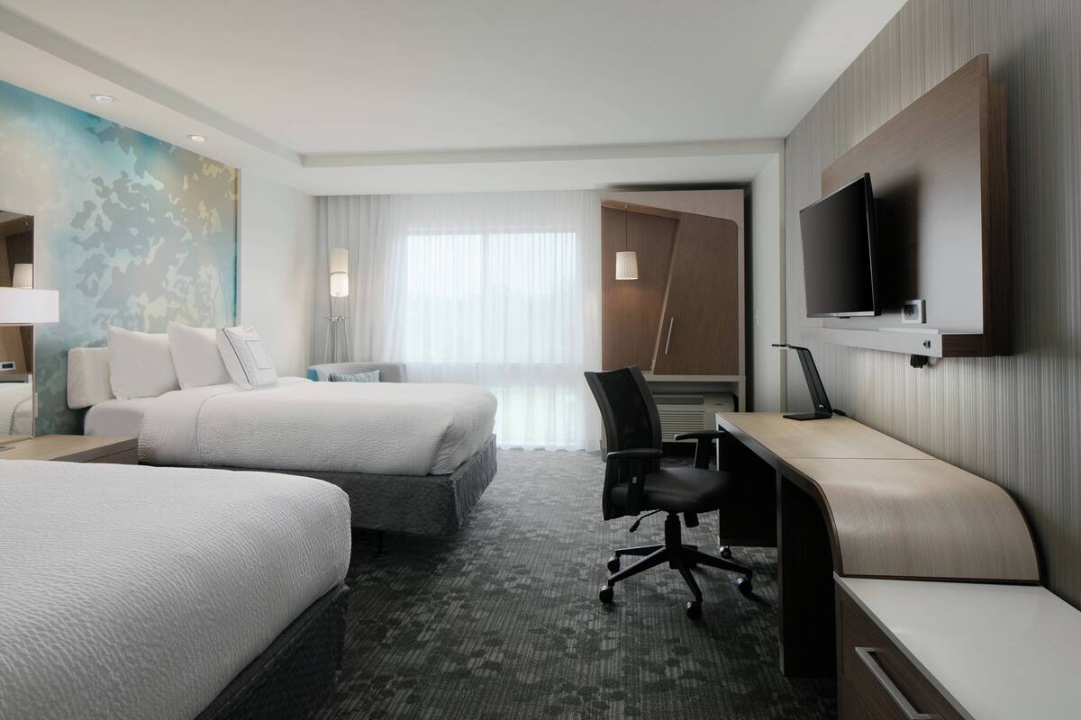 A hotel room with two beds, a desk, a chair, a TV, and a large window with curtains.