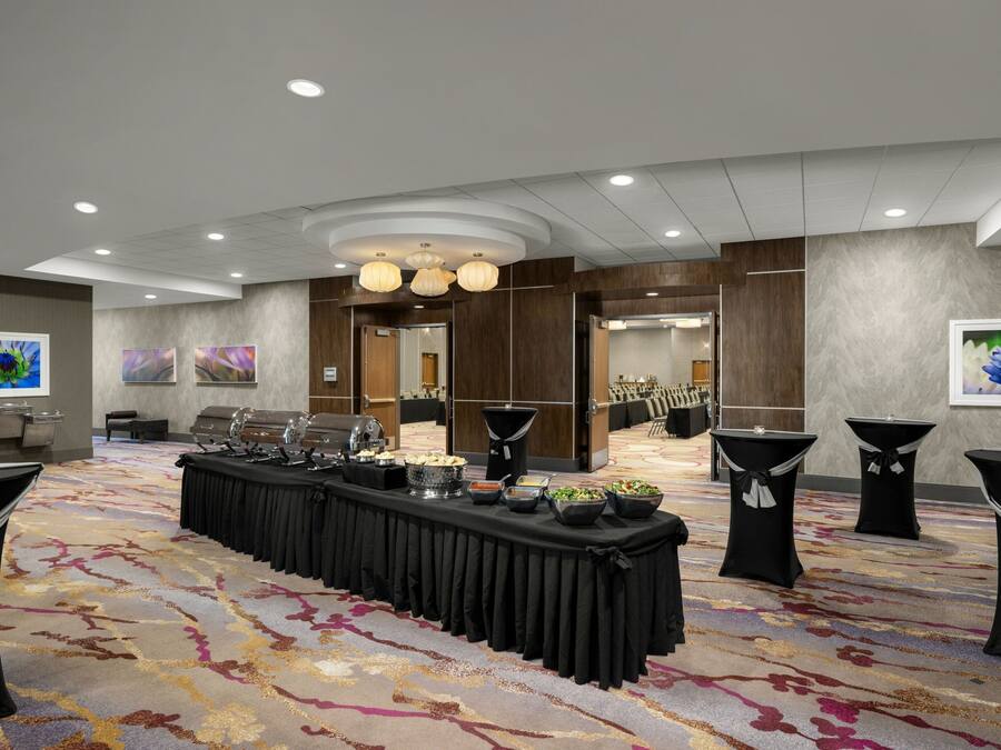 Un salón de banquetes con una larga mesa dispuesta para un bufé, rodeado de sillas y mesas con manteles negros.