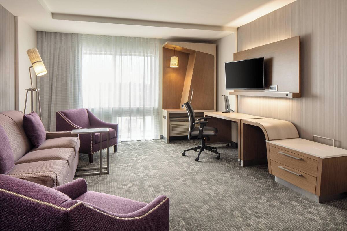 A hotel room with a sofa, a desk, a chair, a television, and a lamp.