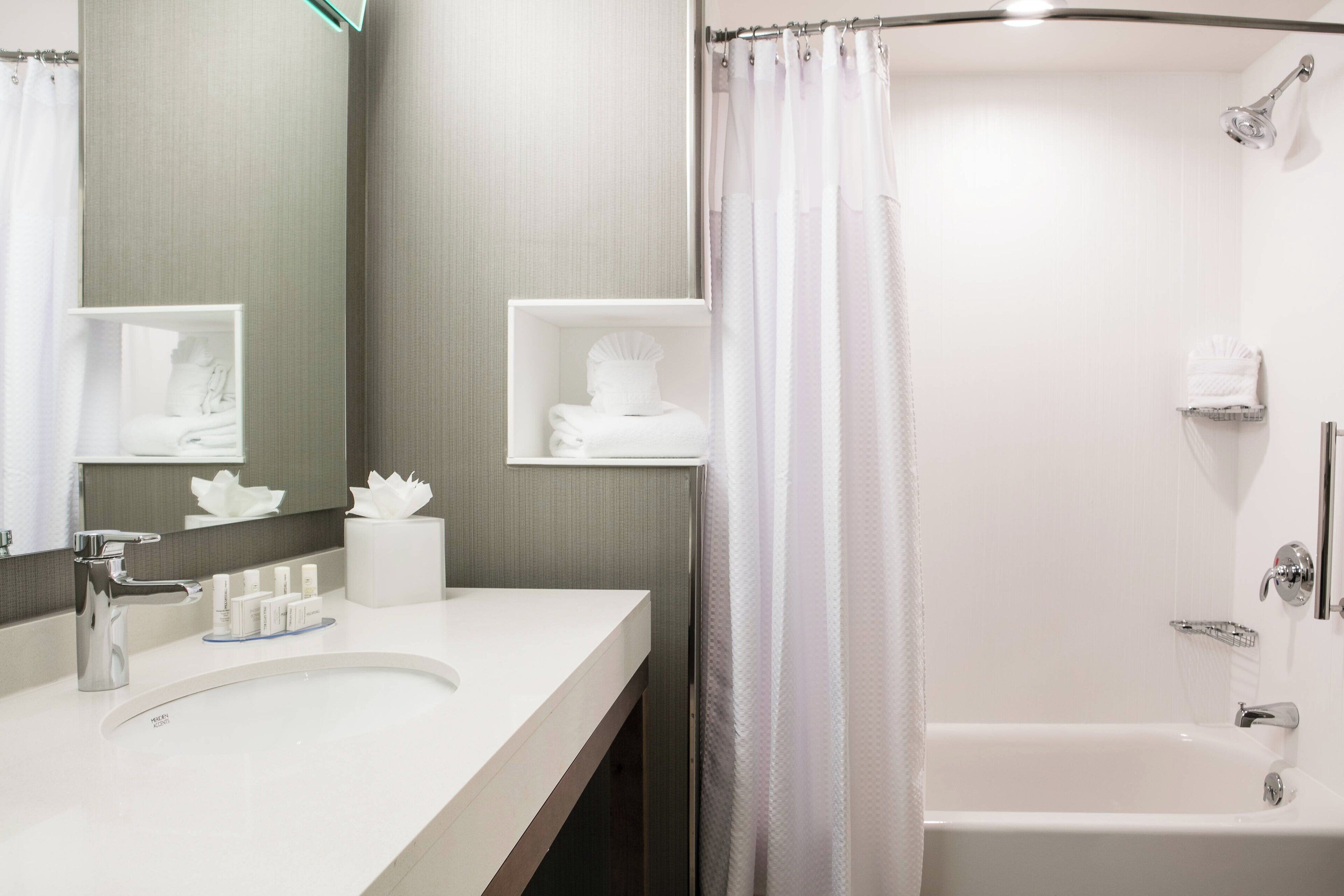 Une salle de bain moderne avec un lavabo blanc, un miroir et une douche. On trouve des articles de toilette sur le lavabo et un porte-serviettes au-dessus du miroir.