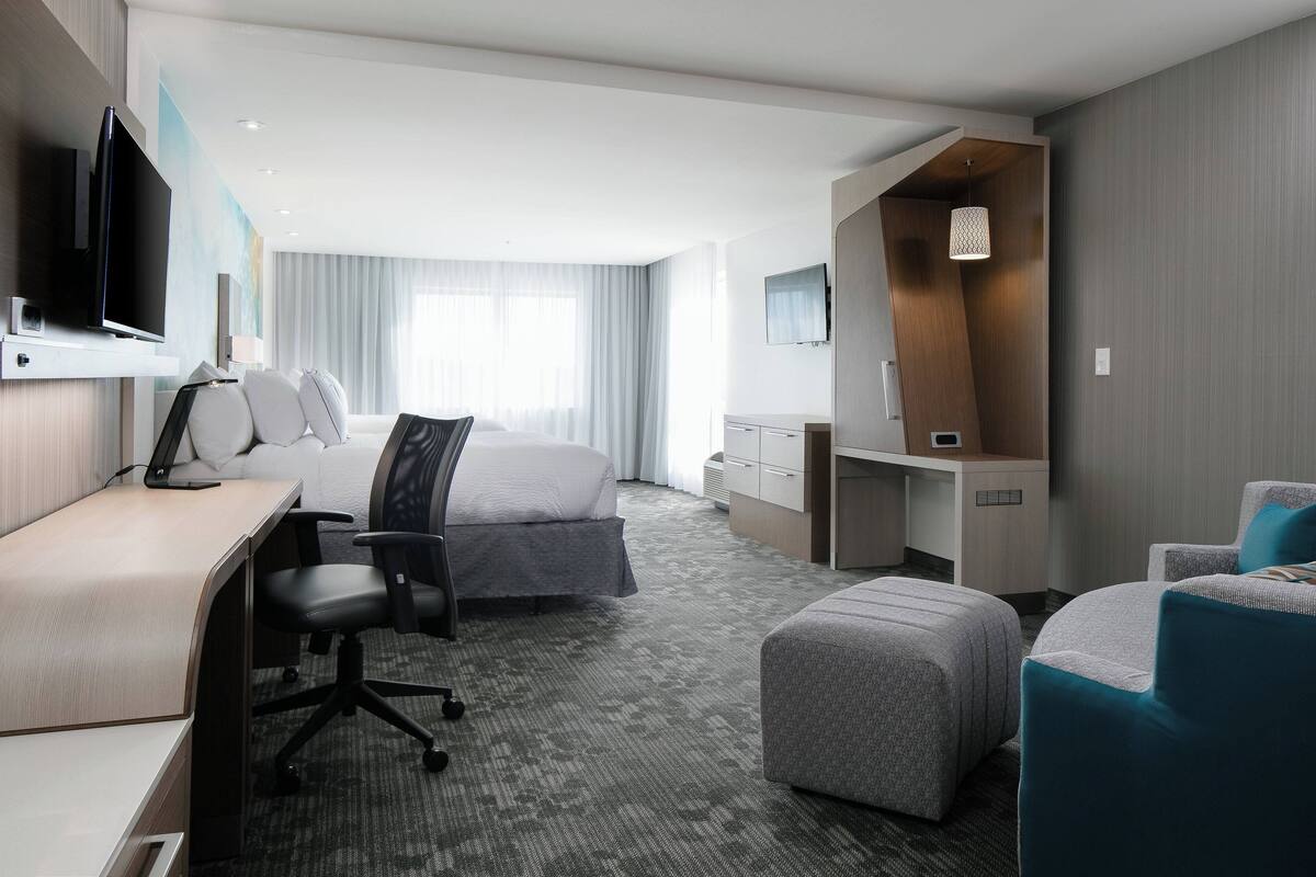 A modern hotel room with a bed, desk, chair, ottoman, and a TV mounted on the wall.