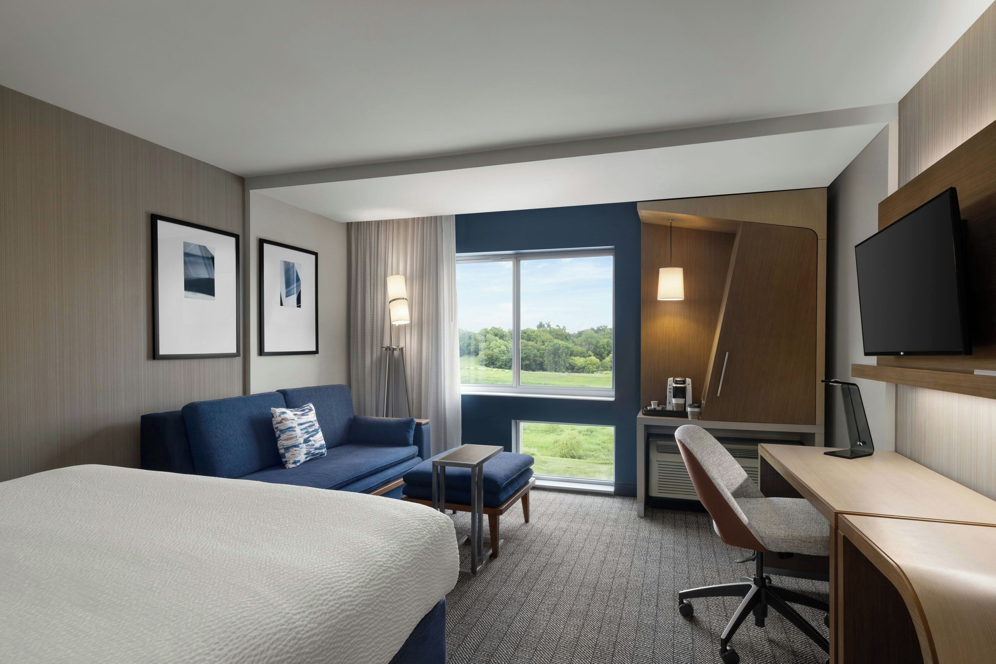 Ein Hotelzimmer mit einem Bett, einer Couch, einem Schreibtisch, einem Sessel, einem Fernseher und einem Fenster mit Blick ins Grüne.