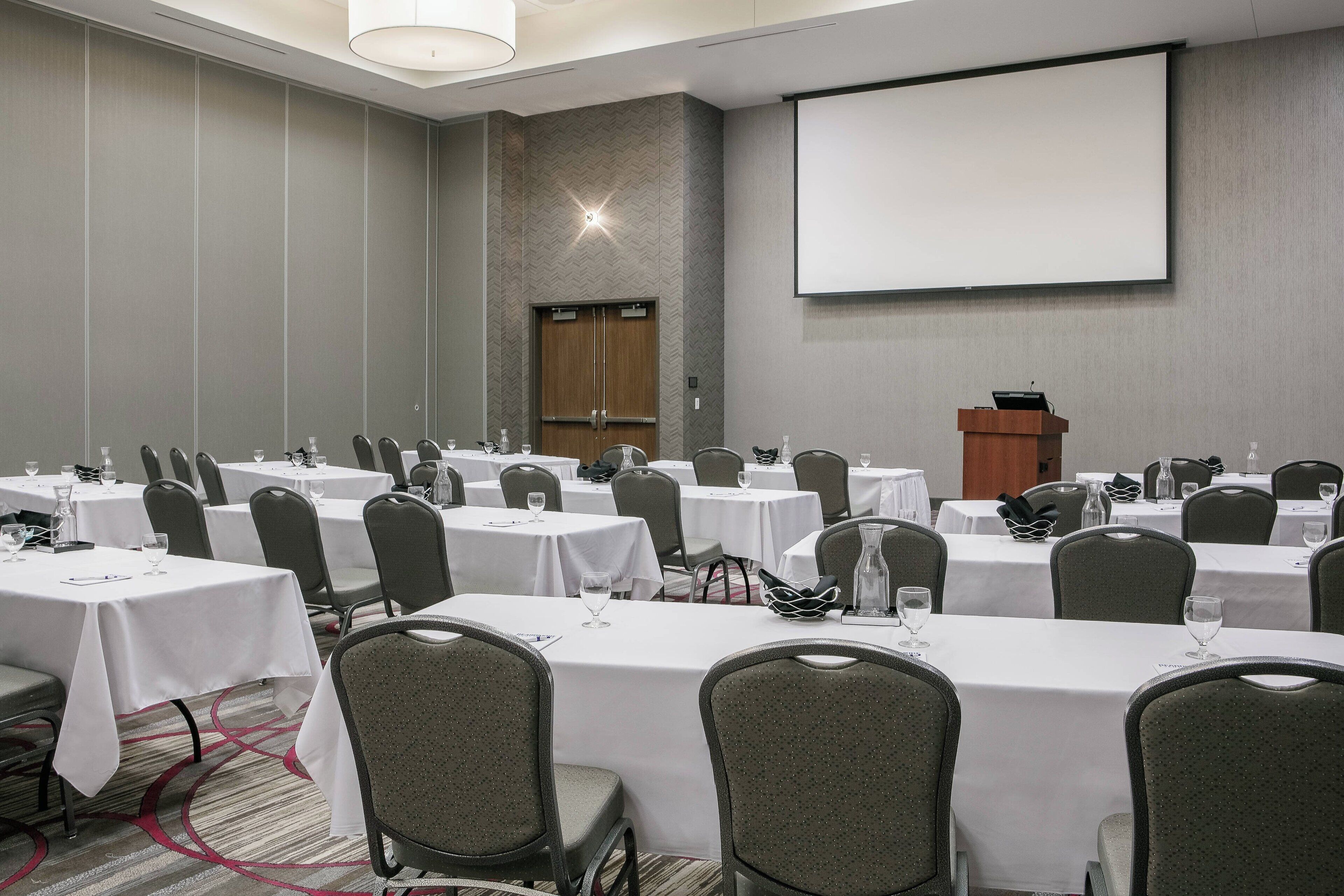Un salón de conferencias con varias mesas, sillas, un atril y una pantalla para proyector.