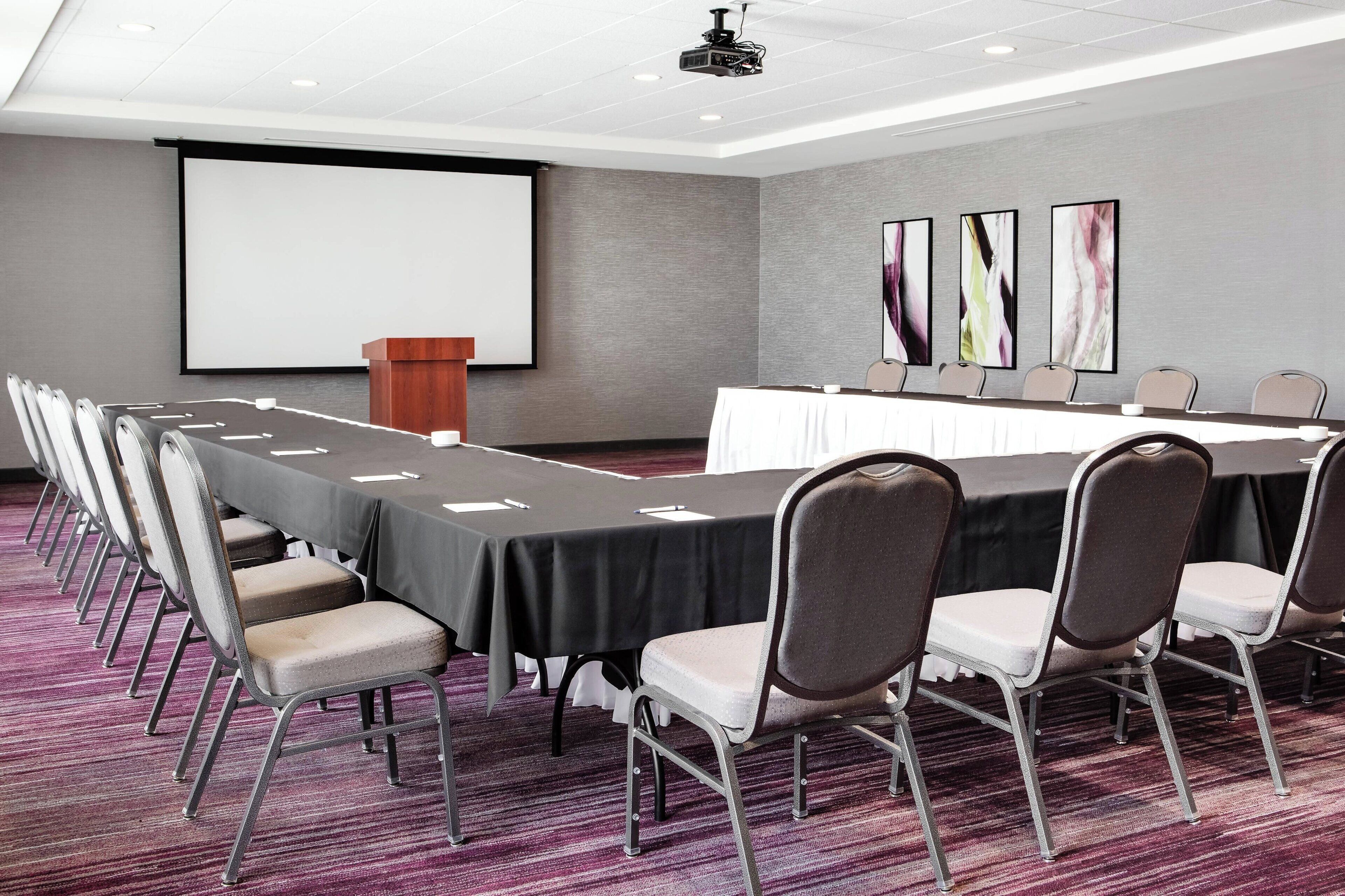 A conference room with a long table, chairs, a podium, and a projector screen.
