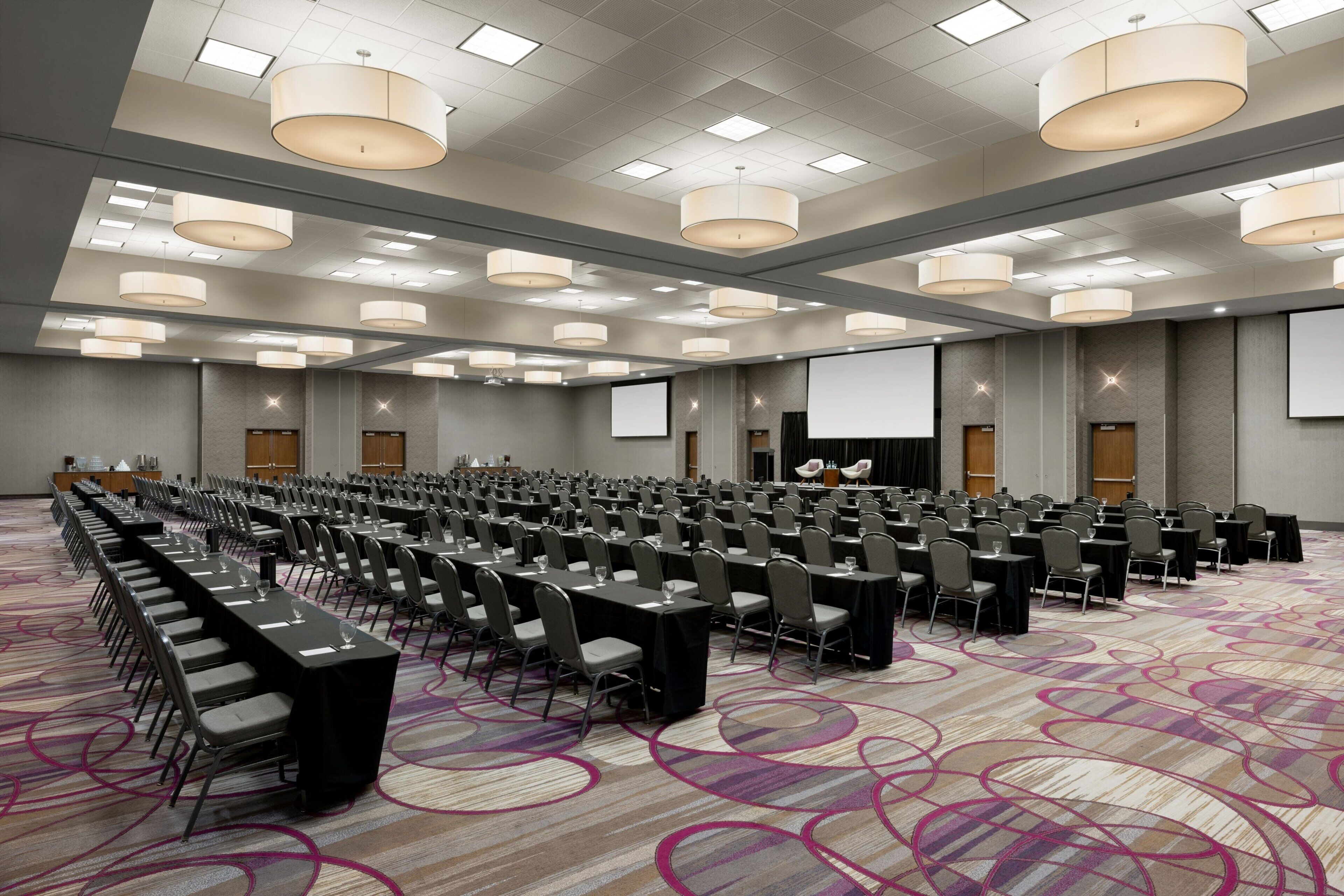 Een ruime congreszaal met rijen tafels en stoelen, een vloerbedekking en in het plafond geïntegreerde verlichting.