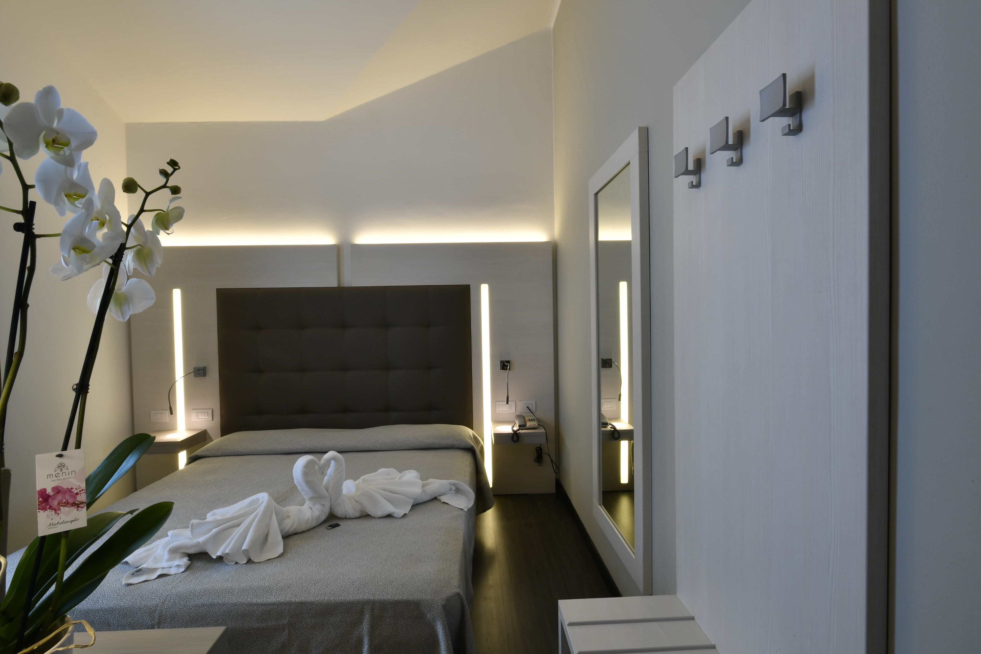Une chambre d’hôtel moderne avec un grand lit, une table de chevet, une commode, un miroir et une plante orchidée en pot.
