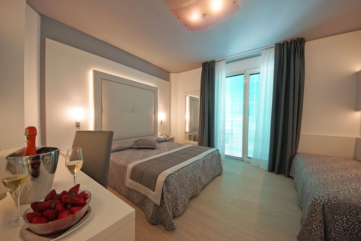 A hotel room with a bed, bedside table with a bottle of champagne and strawberries, a chair, and a view of the outside through a window with curtains.