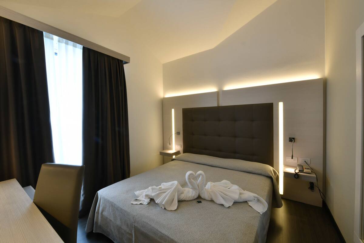 A hotel room with a bed, a desk, a chair, and a nightstand with a lamp.