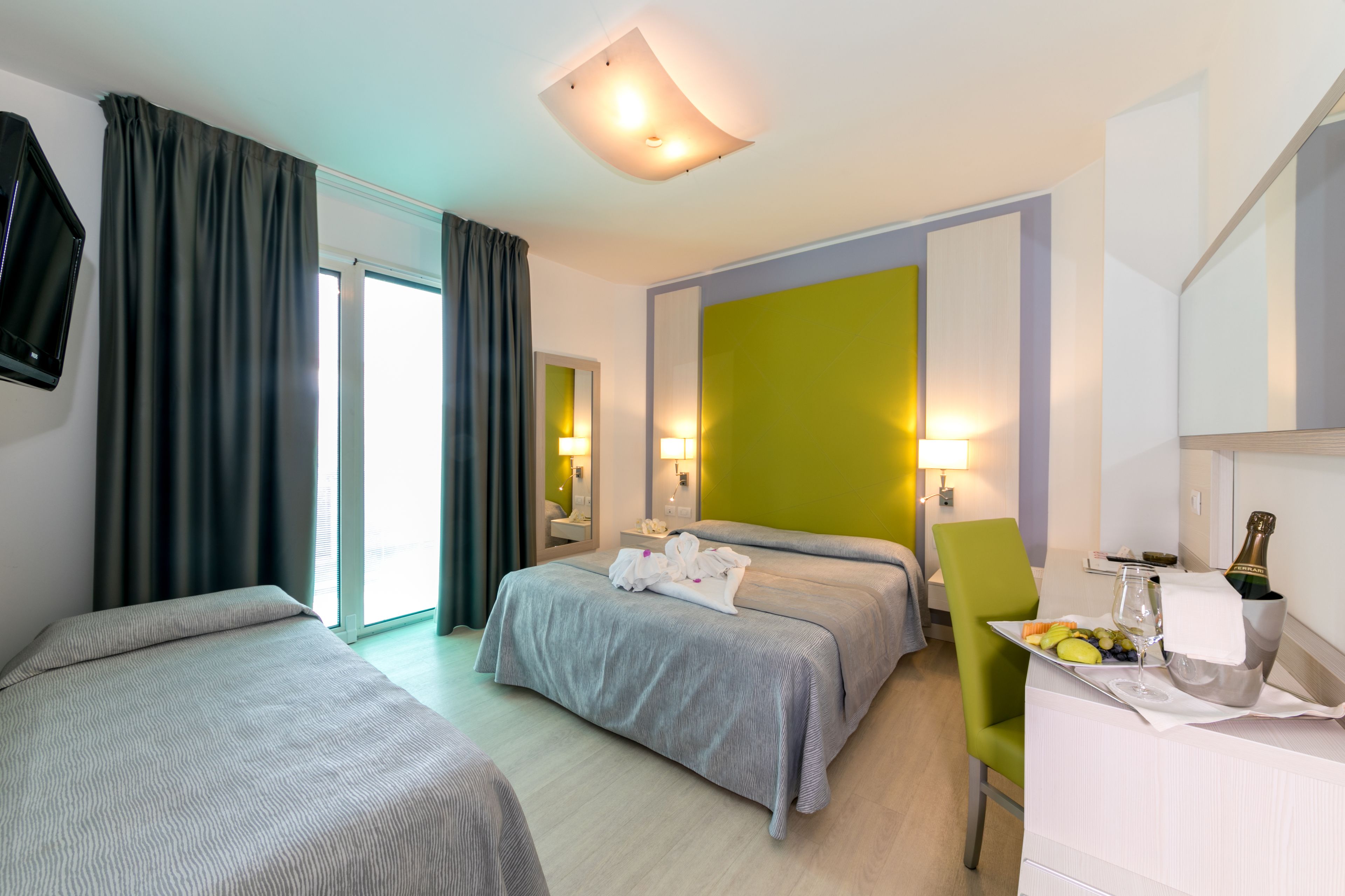 A hotel room with two beds, a green chair, a table with a bottle and glasses, and a large window with curtains.