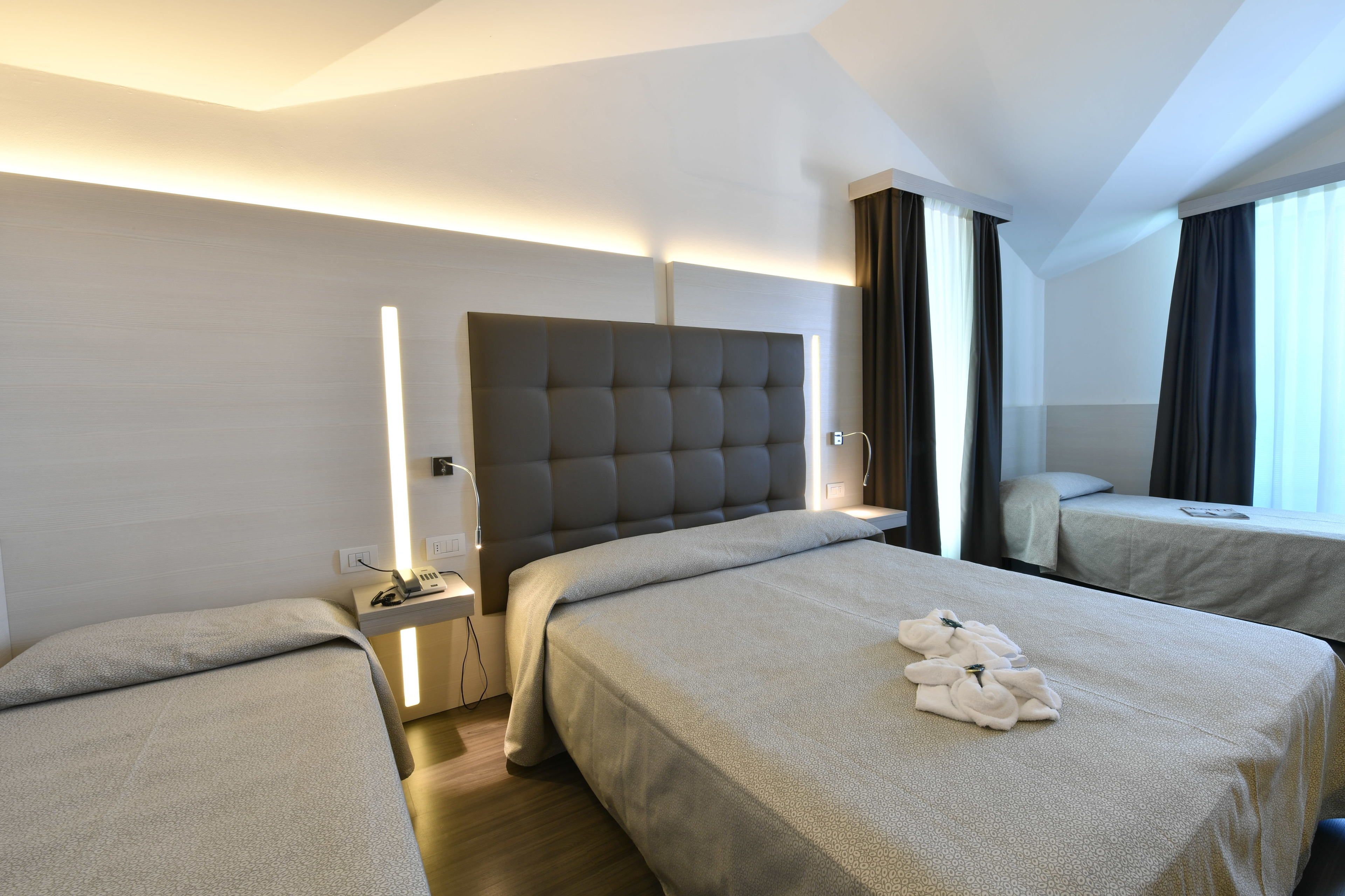A hotel room with two beds, a modern headboard, bedside tables with lamps, and a window with curtains.