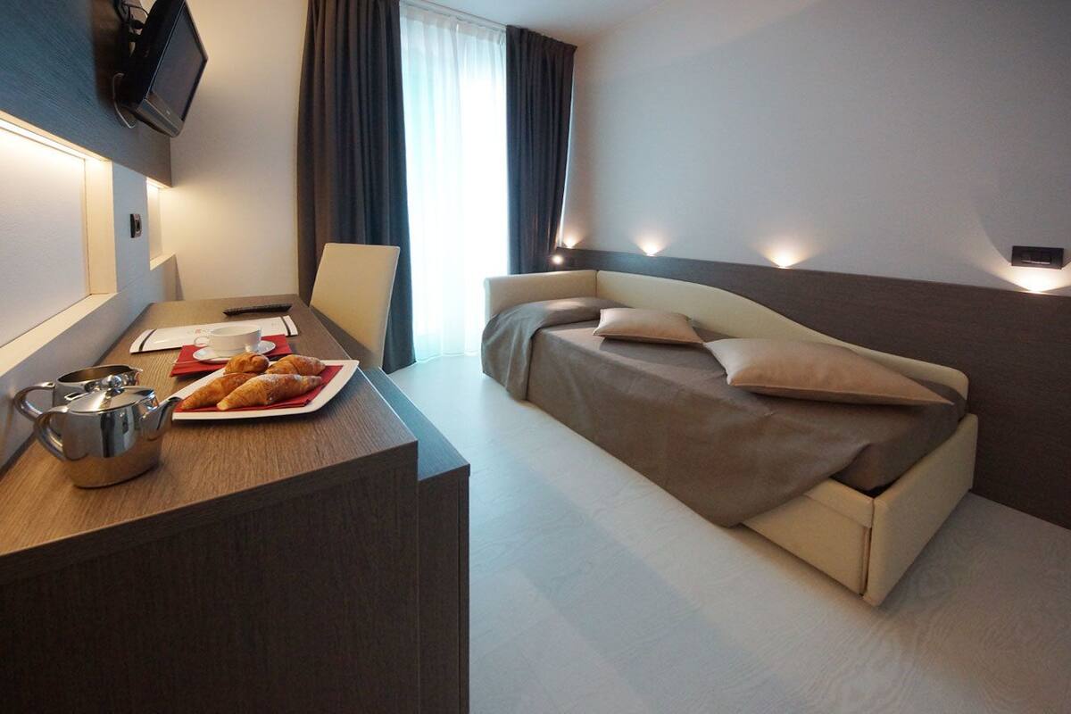 A modern hotel room with a bed, a desk with a teapot and croissants, and a TV mounted on the wall.