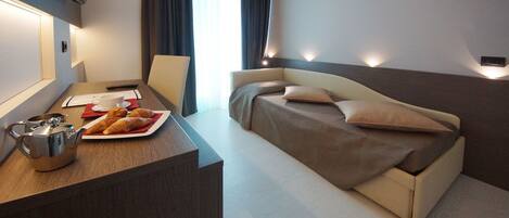 A modern hotel room with a bed, a desk with a teapot and croissants, and a TV mounted on the wall.