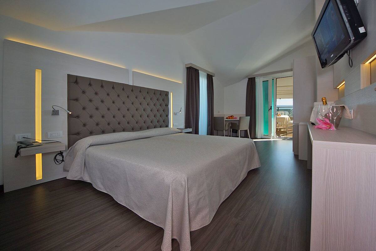 A modern hotel room with a large bed, a flat-screen TV mounted on the wall, and a desk with a lamp.