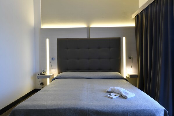 A hotel room with a large bed, a headboard with integrated lighting, bedside tables with lamps, and a dark curtain on the window.