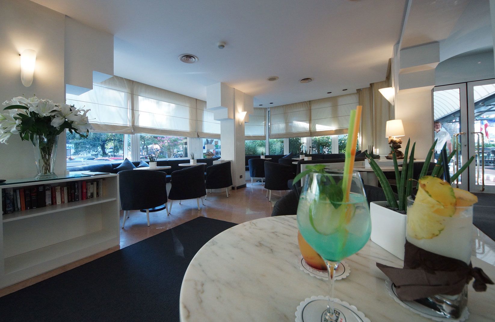 A modern hotel lobby with a marble reception desk, black chairs, a bookshelf with books, and large windows offering a view of the outside.