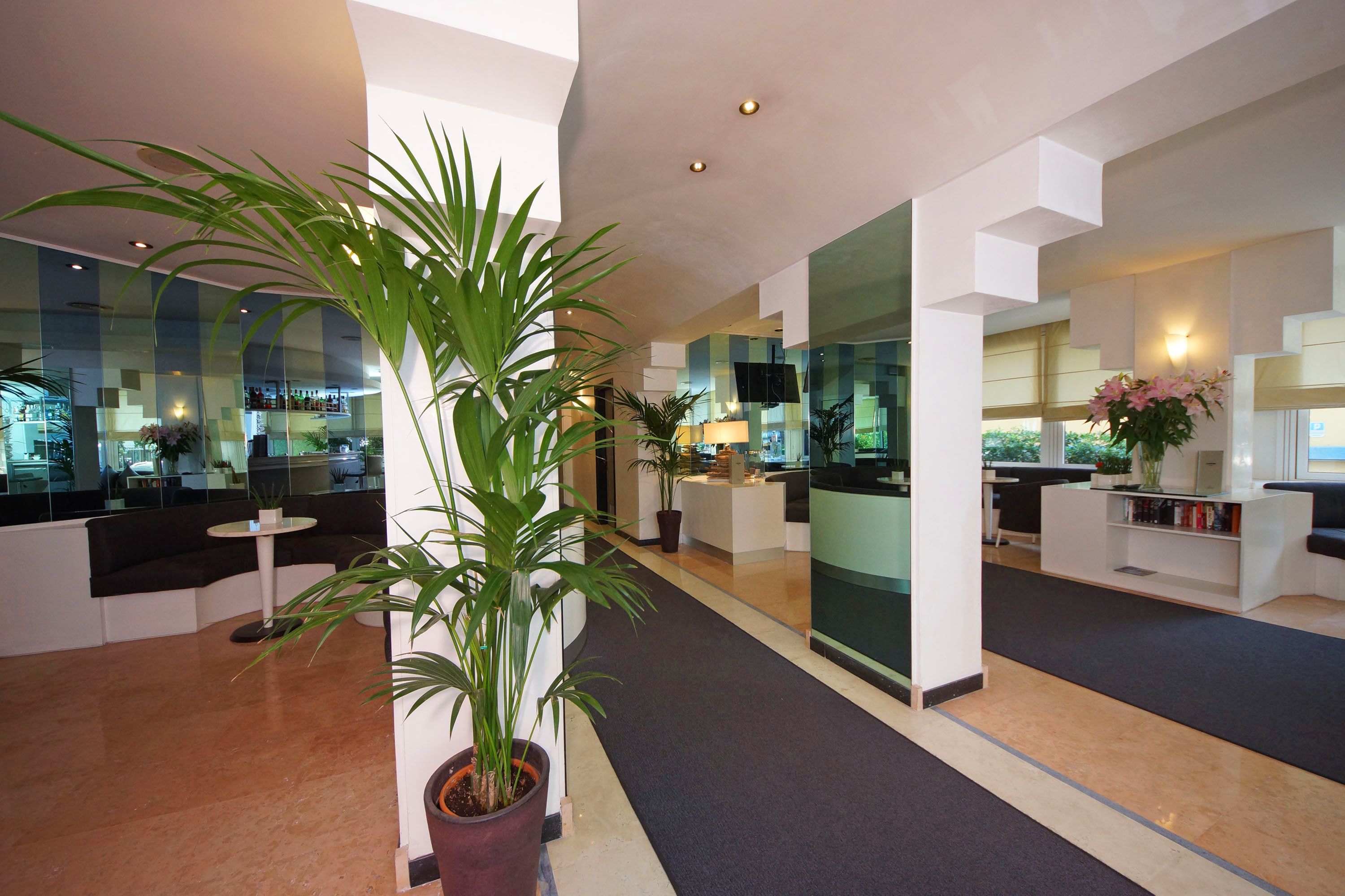 A modern lobby with a central potted plant, a reception desk, and a glass wall reflecting the interior.
