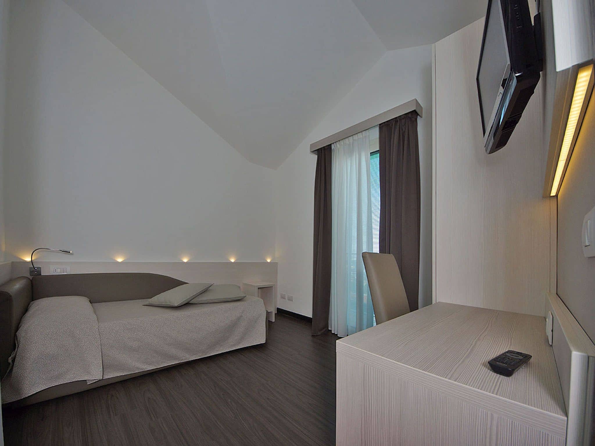 Una habitación de hotel moderna con una cama, un escritorio, una silla, un televisor montado en la pared y una ventana con cortinas.