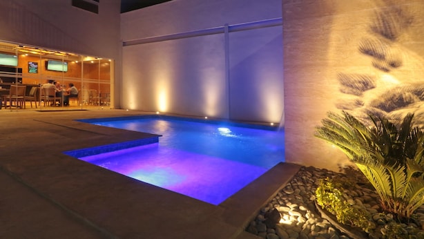 A swimming pool with a blue-lit edge, a bar area with people, and a wall-mounted TV. - Bluu Hotel (Frontera)