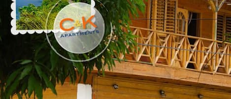 A wooden balcony with a railing, a sign for C&K Apartments, and some greenery.