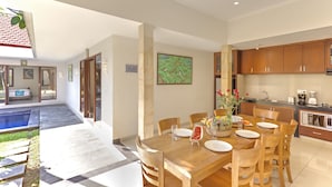 A spacious dining area with a wooden table set for four, a kitchen with wooden cabinets, and a view of a swimming pool. - Villa Kedis (Canggu)