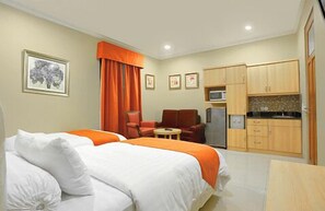 A hotel room with two beds, a small dining table, a kitchen area with cabinets and appliances, and a red chair. - Rumah Aca (Bandung)