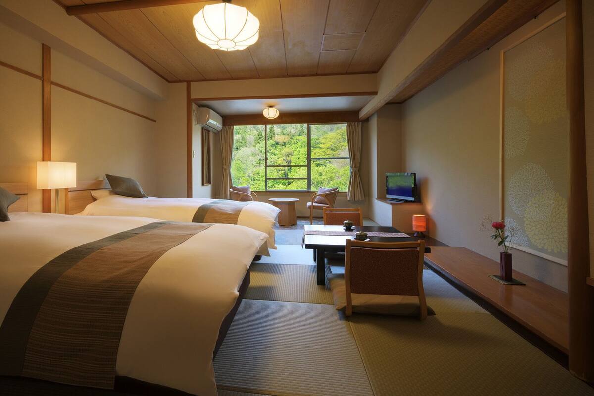 A hotel room with two beds, a dining table, chairs, a TV, and a window with a view of greenery.