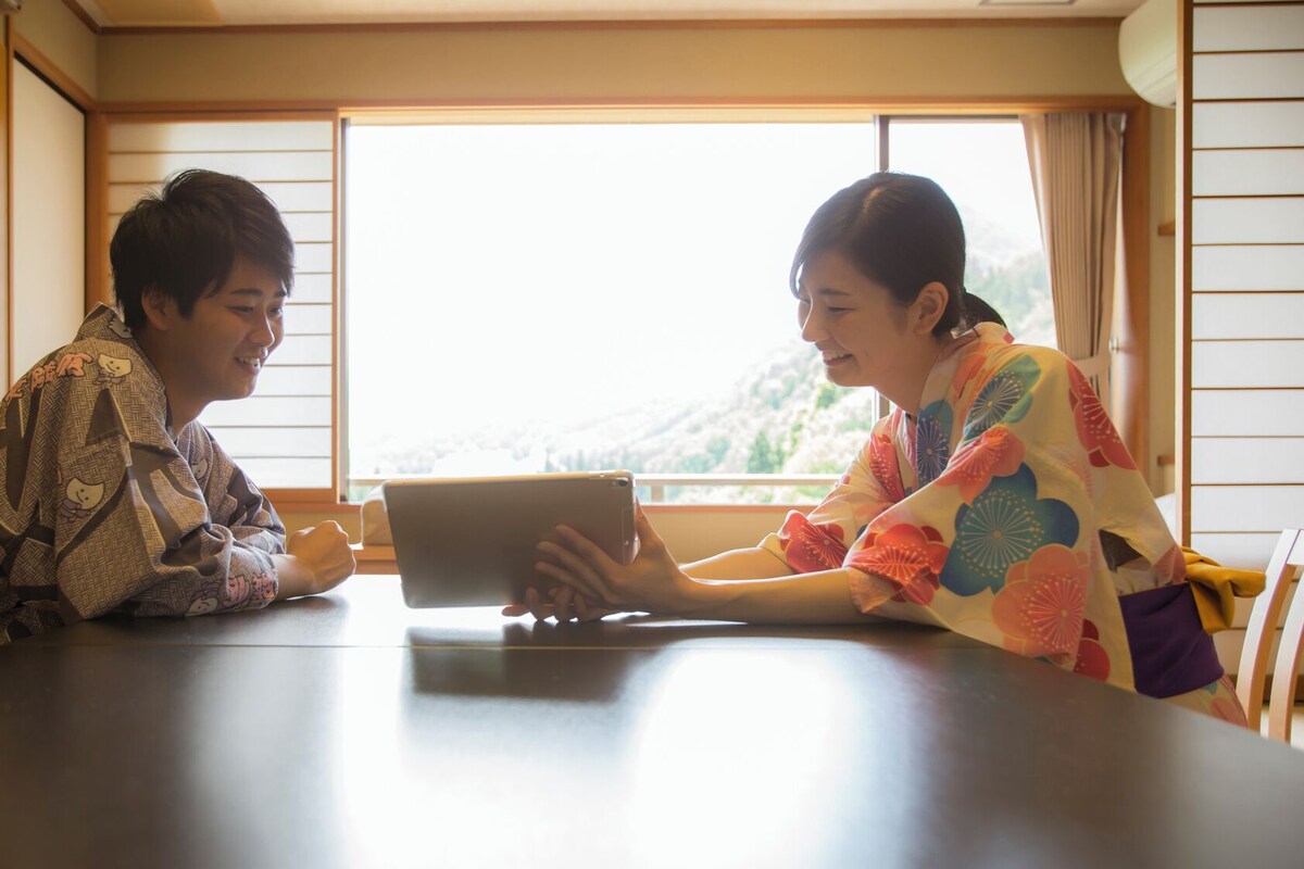 Two people sitting at a table with a tablet, in a room with a view of a mountain through a window.