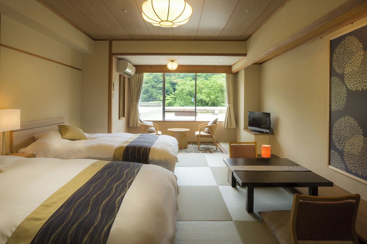 A hotel room with two beds, a TV, a table, and a view of greenery outside.