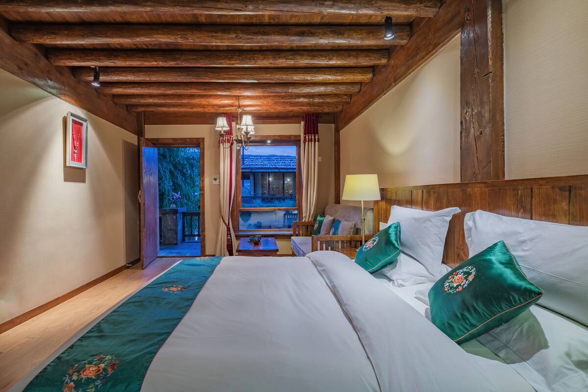 Une chambre d’hôtel avec un plafond en bois, un grand lit avec du linge de lit blanc et des oreillers verts, une table de chevet avec une lampe, et une vue sur un balcon avec des rideaux.