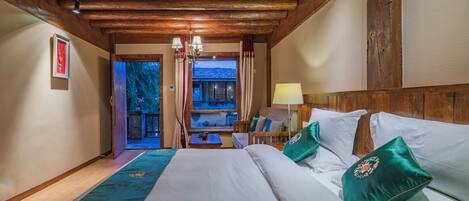 A hotel room with a wooden ceiling, a large bed with white linens and green pillows, a nightstand with a lamp, and a view of a balcony with curtains.