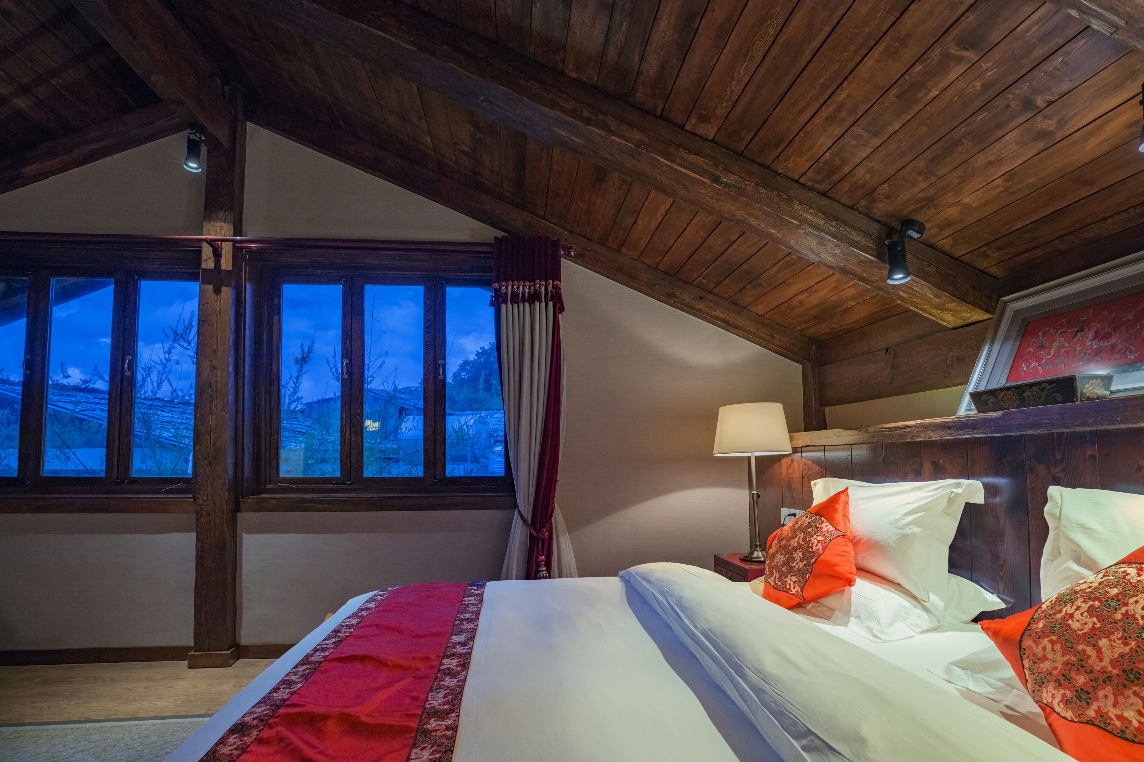 A bedroom with a wooden ceiling, a large window with curtains, a bed with white and orange bedding, and a nightstand with a lamp.