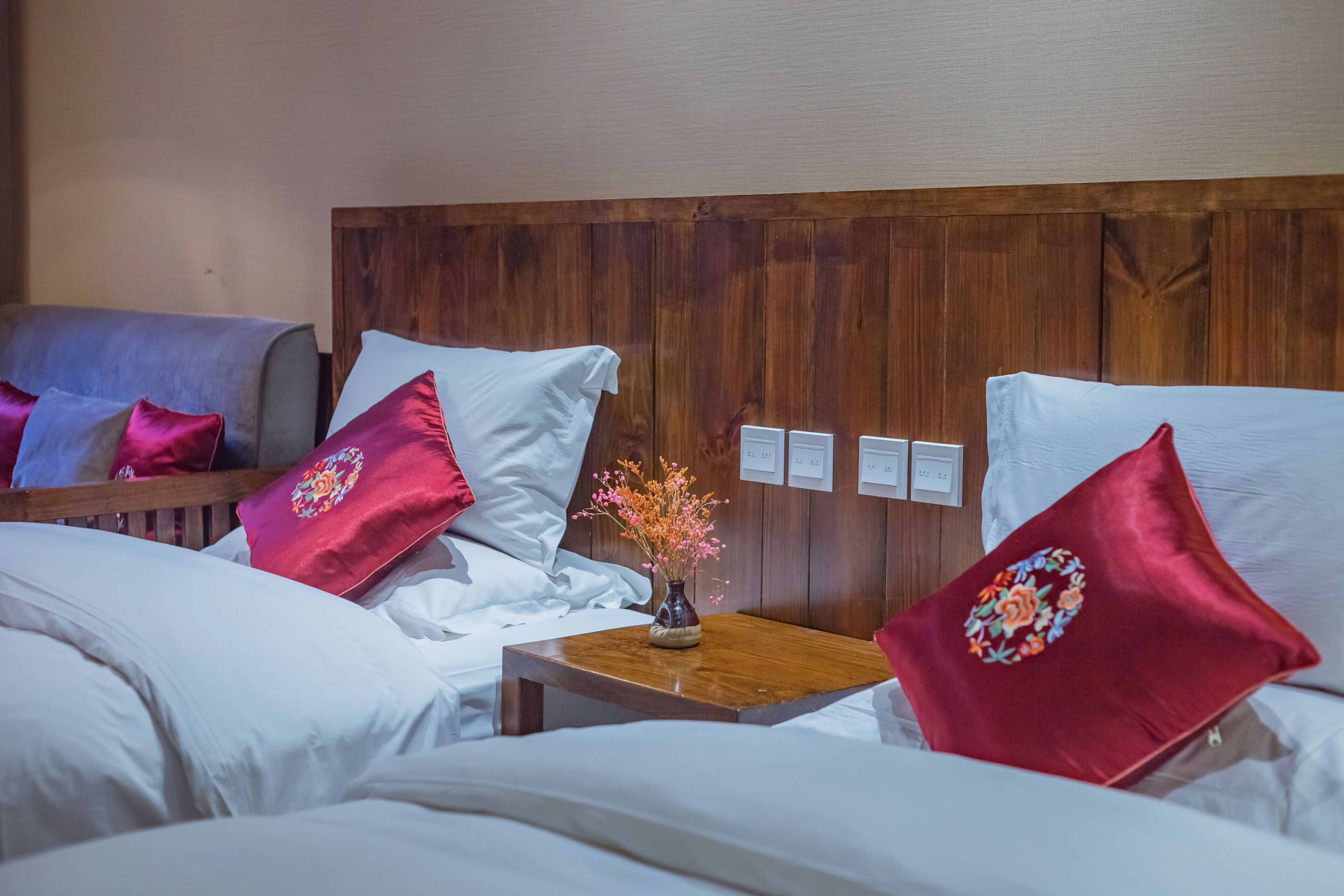 A hotel room with two beds, each with white linens and red pillows. There is a small wooden table with a vase of flowers between the beds.