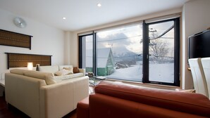 A modern living room with a sofa, a coffee table, and a large window offering a view of a snowy landscape and a green-roofed building. - Holiday Niseko Cecuko (Kutchan)