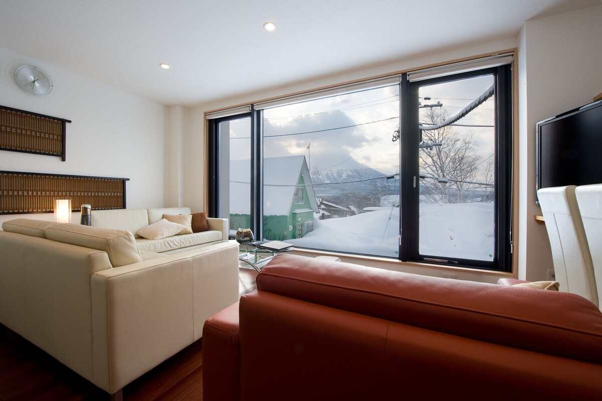 A modern living room with a sofa, a coffee table, and a large window offering a view of a snowy landscape and a green-roofed building.