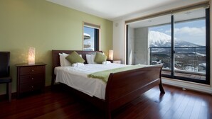 A bedroom with a large bed, a nightstand, a lamp, and a view of a snowy mountain through a large window. - Holiday Niseko Cecuko (Kutchan)