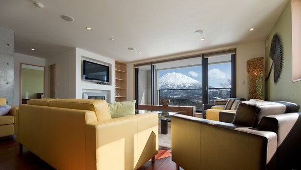 A modern living room with a sofa, armchairs, and a TV. A large window offers a view of a mountain. - Holiday Niseko Cecuko (Kutchan)