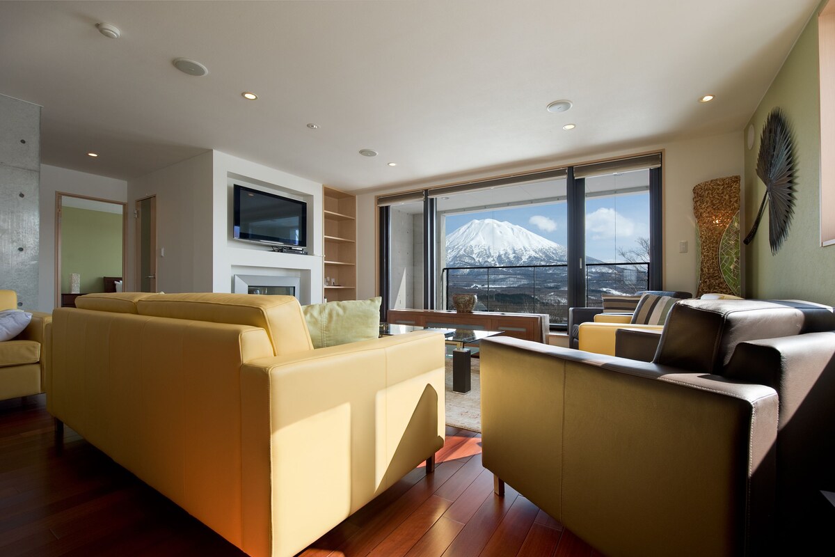 A modern living room with a sofa, armchairs, and a TV. A large window offers a view of a mountain.