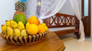 Una cesta de frutas sobre una mesa de madera, con una cama y mosquitera al fondo.