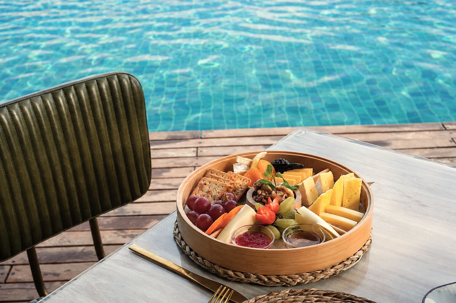A wooden fruit and cheese board with a pool in the background.