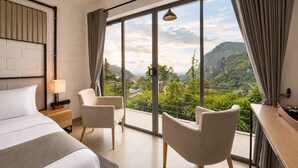 A modern hotel room with a large bed, two armchairs, a nightstand, a lamp, and a view of a mountainous landscape through floor-to-ceiling windows. - Onrock Boutique Hotel (Ella)