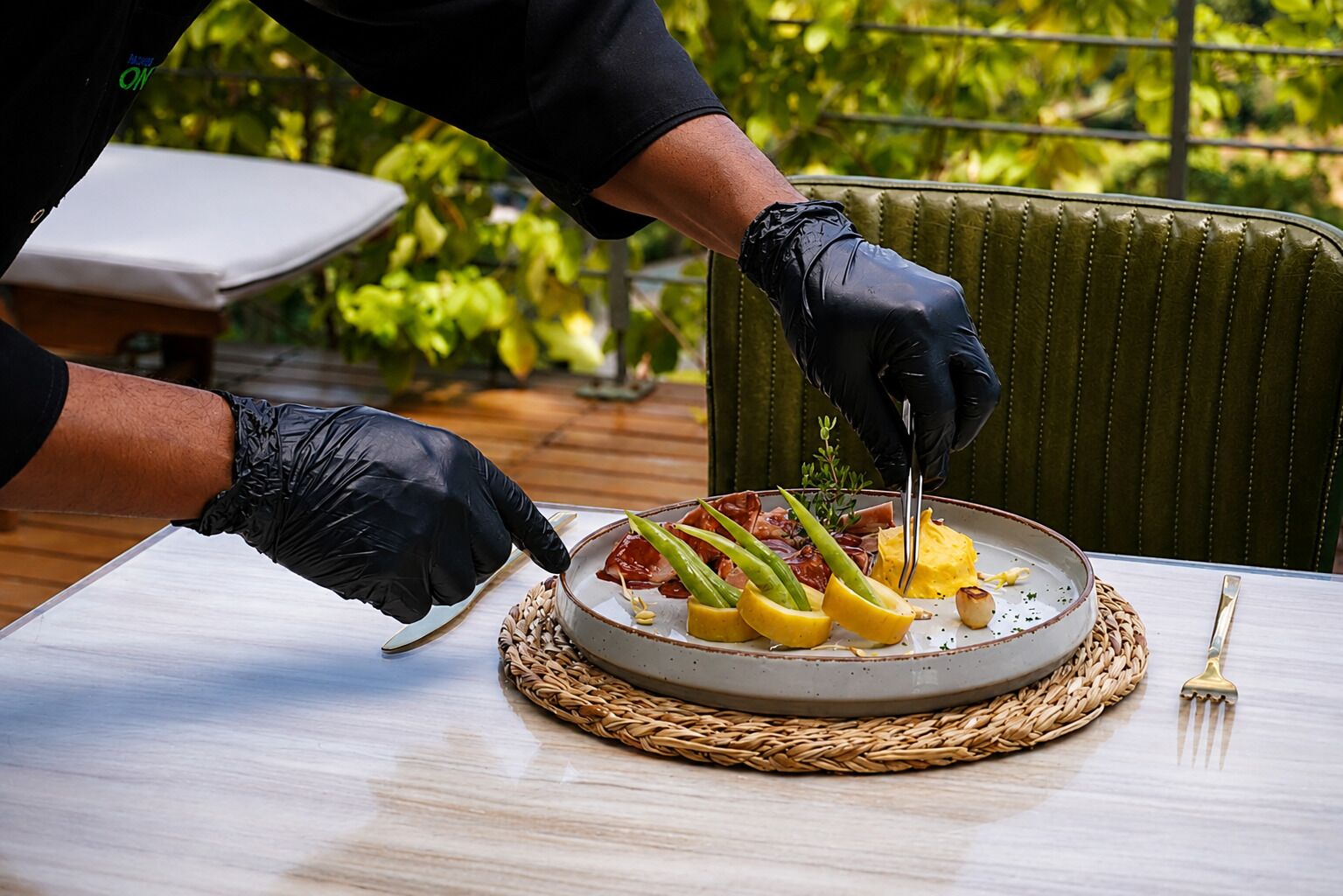 A person serving food on a table with a woven placemat, a chair, and greenery in the background.