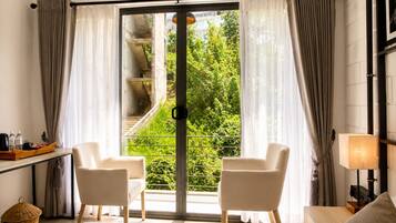 Habitación de hotel con un gran ventanal corredero, dos sillas, una cama, una mesita de noche, una lámpara y vistas a zonas verdes y una escalera.