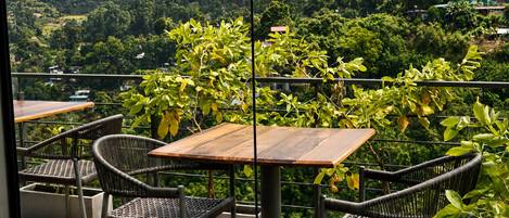 A balcony with wicker chairs and a wooden table, overlooking a lush green landscape with hills in the background.