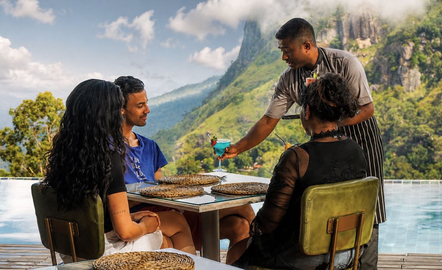 A dining table with woven placemats, a person serving a drink, and a mountainous landscape in the background.