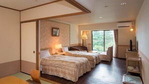 A hotel room with two beds, a small table, a chair, a TV, and a balcony with a view of greenery.