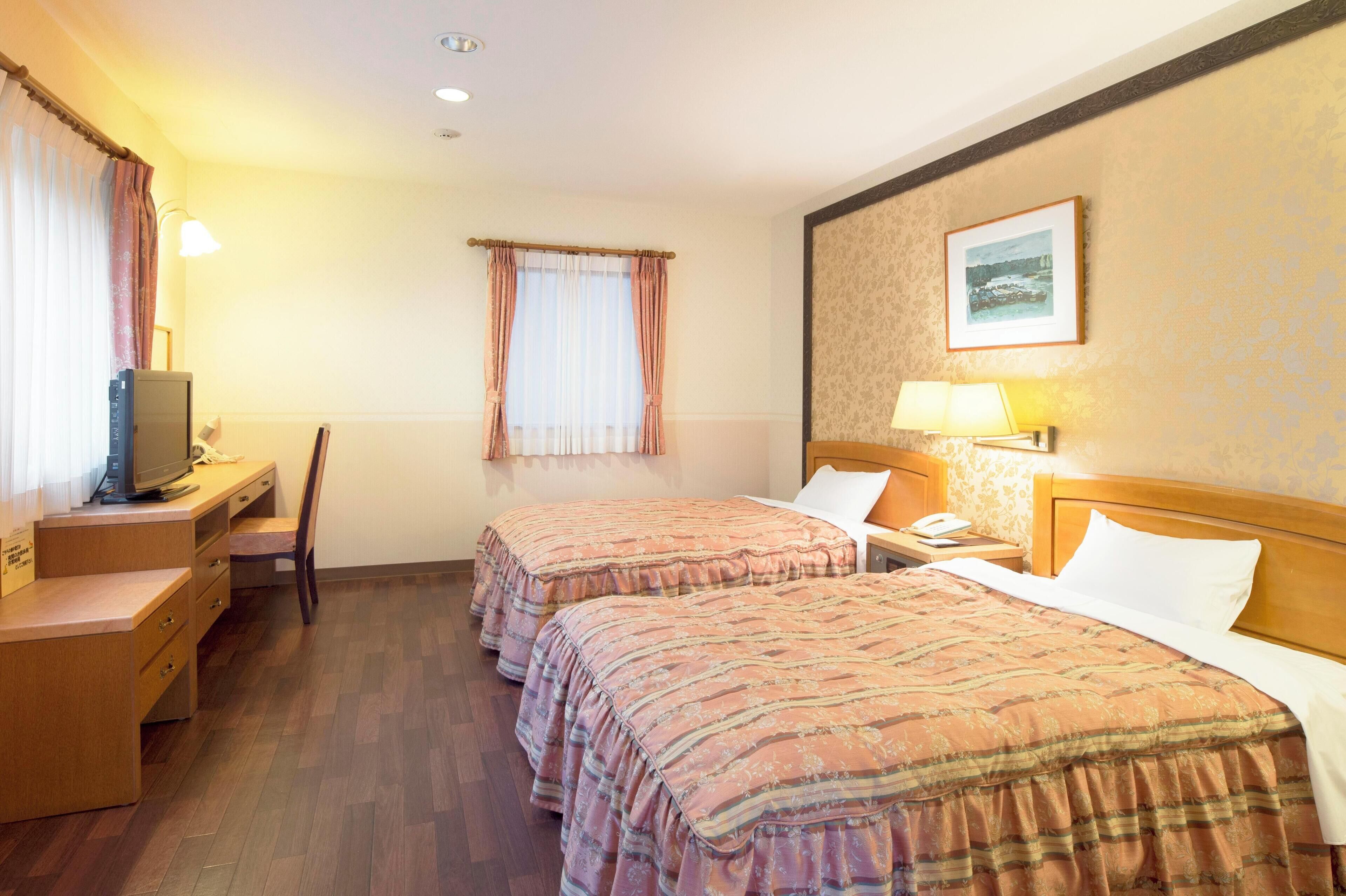 Una camera d'albergo con due letti, una scrivania con un computer, una sedia, un comodino con una lampada e una finestra con tende.