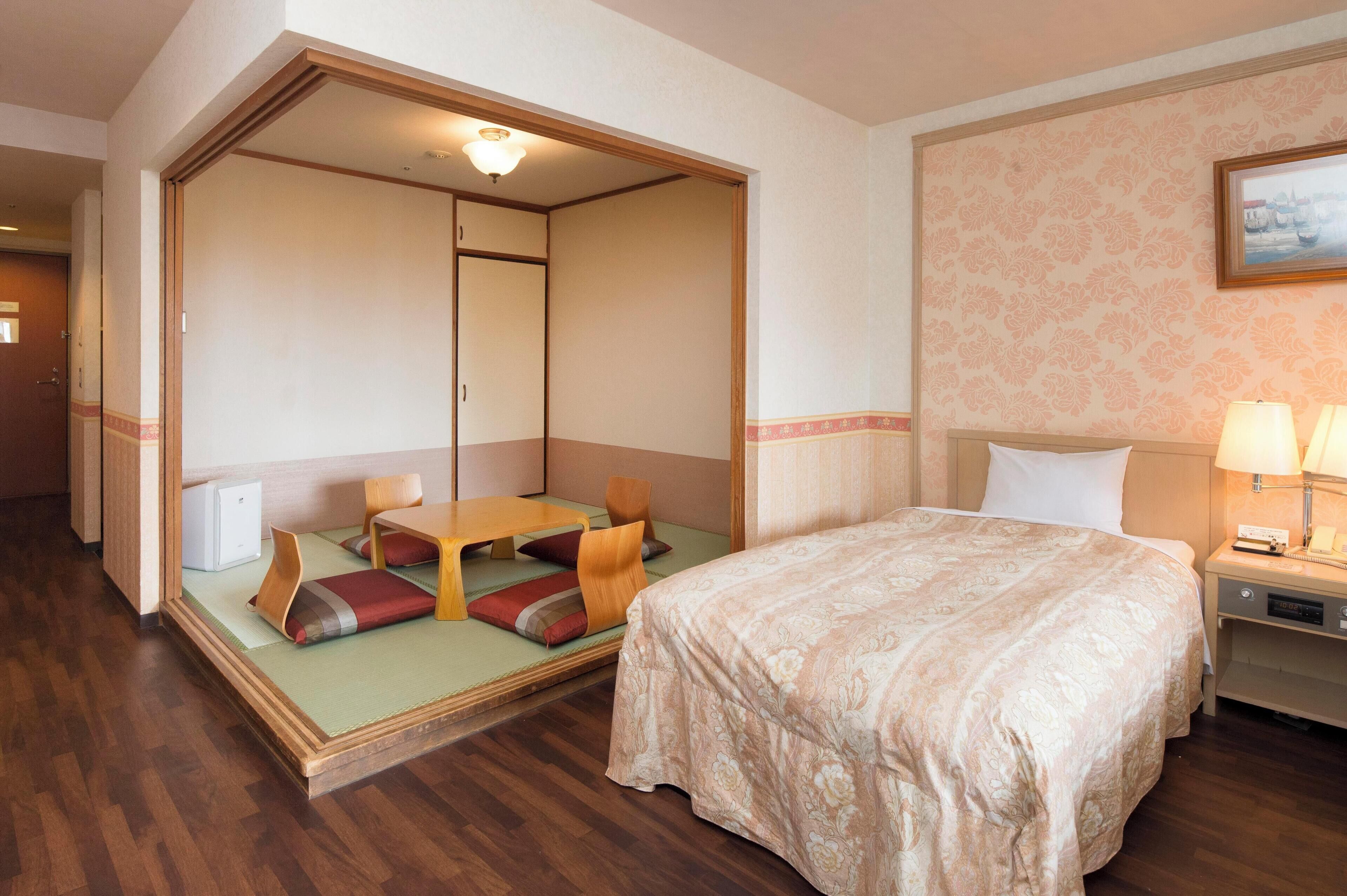A hotel room with a bed, a small table with chairs, a futon bed, and a nightstand with a lamp.